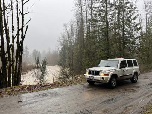 A white SUV is parked on a wet road with river behind it. The sky is grey with leafless trees.