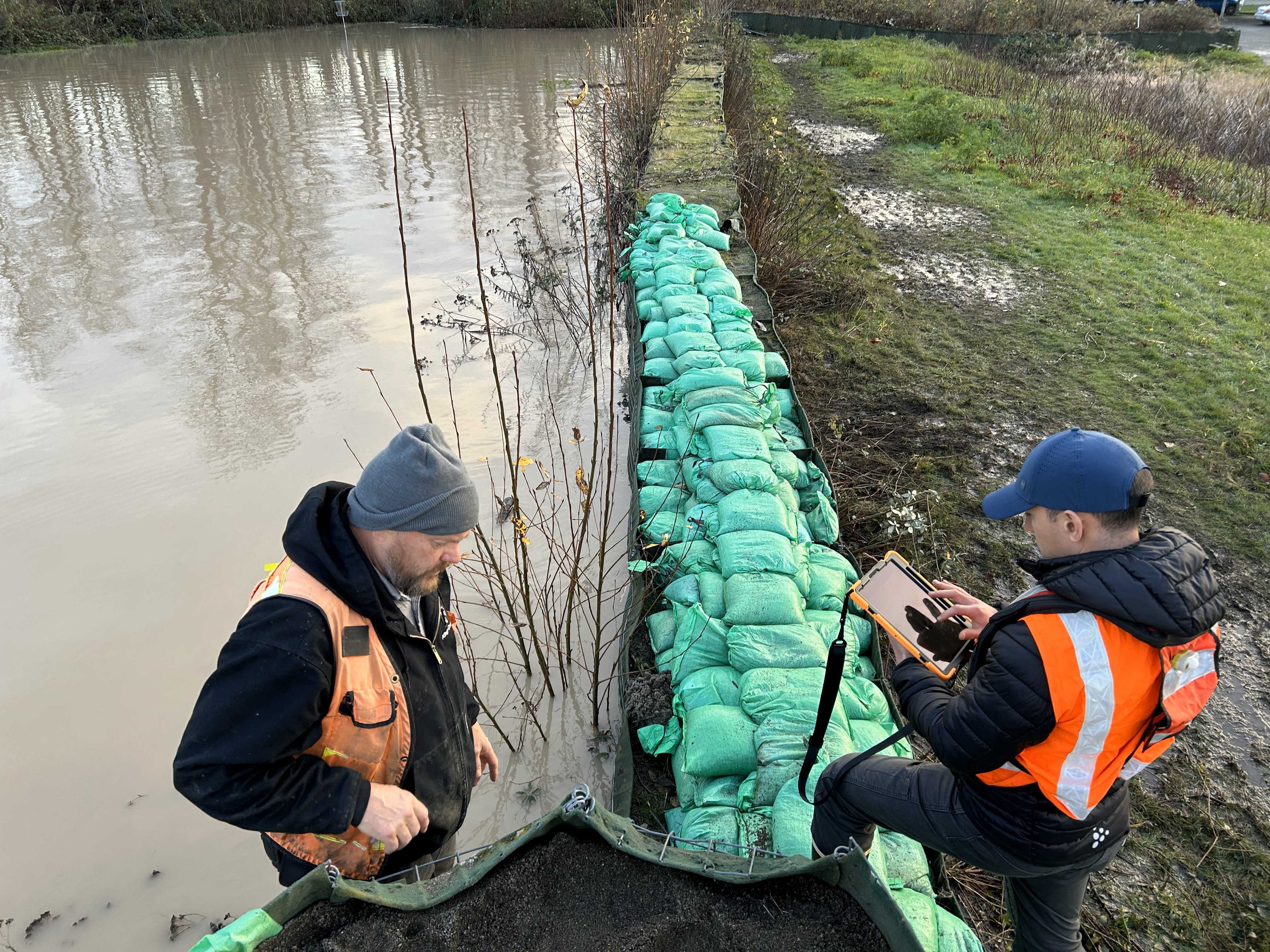 Two workers in safety vests inspect green sandbags reinforcing a flooded riverbank; one stands in shallow water while the other uses a tablet.