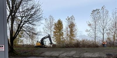 A bulldozer atop a hill that comes from a parking lot. Two people in personal protective equipment stand to the far right on top of the hill.