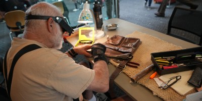 A person with a visor and glasses sits at a table repairing a leather bag.