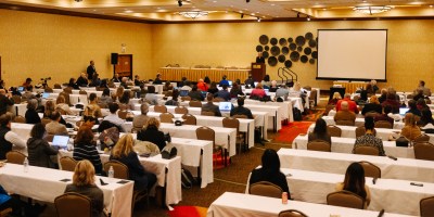 Large conference room filled with attendees seated at long tables, facing a stage where a panel of speakers presents alongside a projection screen.