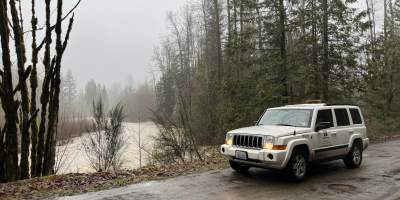 A white SUV is parked on a wet road with river behind it. The sky is grey with leafless trees.