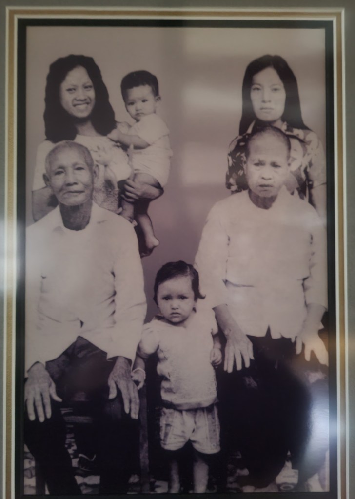 Black and white photo of a family. •	BabyMay is the only baby that survived the war. Maythia’s mom is the woman on the left and she is holding baby Maythia. Maythia’s grandparents are sitting in the photo and the other child his her cousin. 