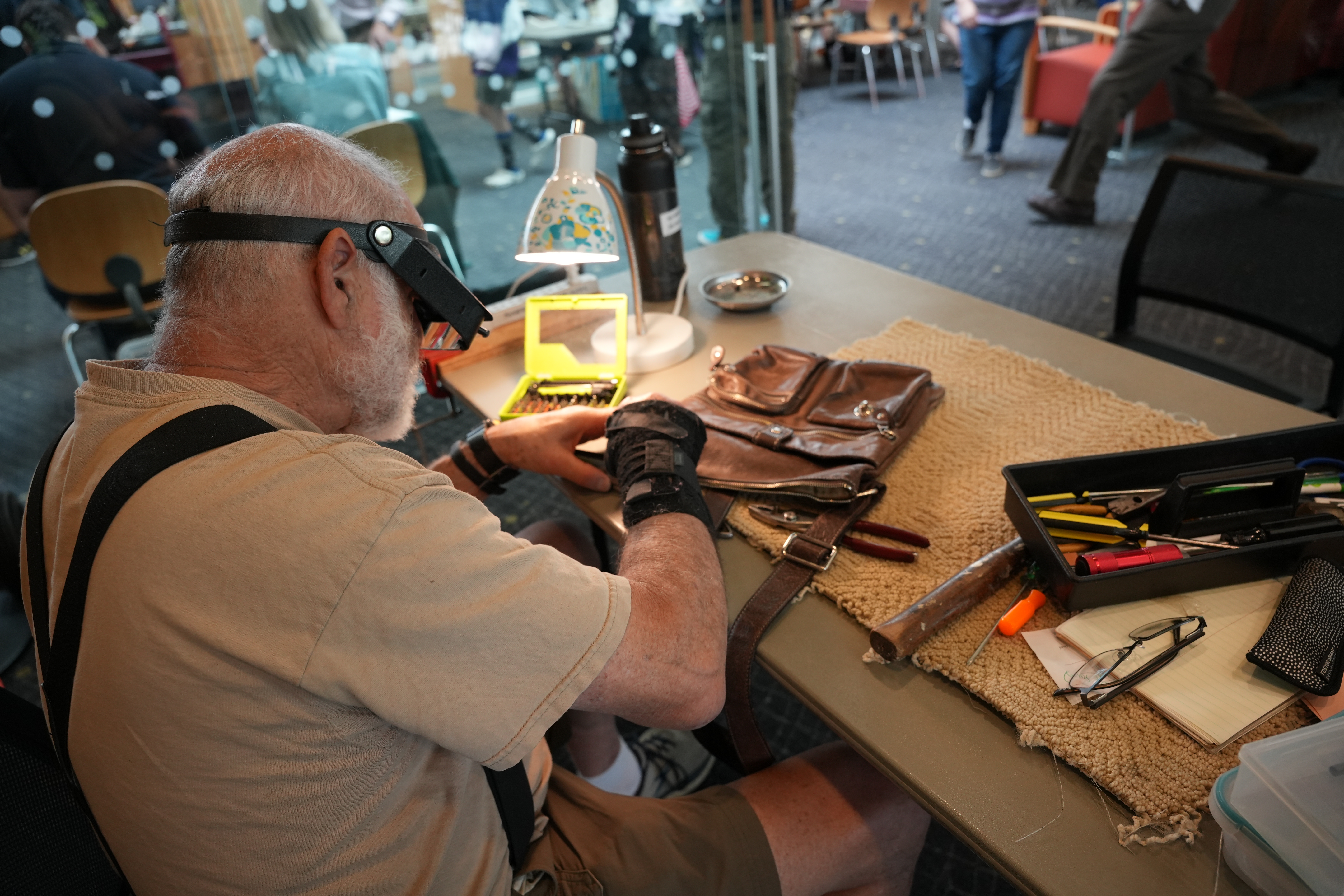 A person with a visor and glasses sits at a table repairing a leather bag.
