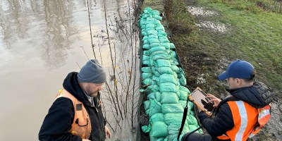 Two workers in safety vests inspect green sandbags reinforcing a flooded riverbank; one stands in shallow water while the other uses a tablet.