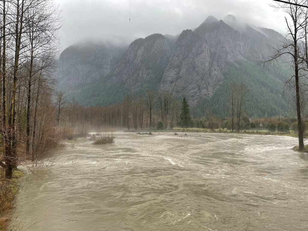 View the side of the North Fork Snoqualmie. The mountain ridge is in the background and has some clouds at the top. Trees along the river mostly have no leaves, but there are some evergreens in the distance.