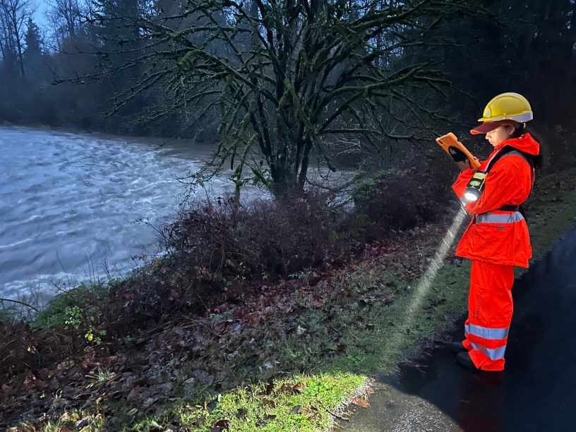 A person wearing an orange safety suit, hardhat, holds a flashlight and enters data on a screen on the banks of the White River during a flood event.