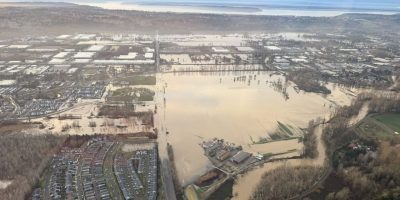 View of the Green River from a helicopter. Brown floodwater extends over one side of the riverbank covering a major road near a neighborhood and low-lying areas. Puget Sound and the Olympic Mountains are on the horizon.