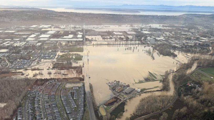 View of the Green River from a helicopter. Brown floodwater extends over one side of the riverbank covering a major road near a neighborhood and low-lying areas. Puget Sound and the Olympic Mountains are on the horizon.
